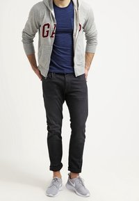 Felpa grigia con zip con logo "GAP", t-shirt blu navy, jeans neri e scarpe da ginnastica grigie. Outfit casual con texture sovrapposte.