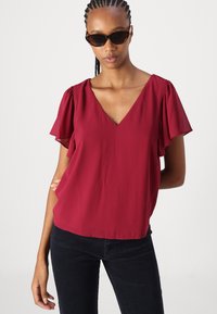 Burgundy blouse with a V-neck, short flutter sleeves, and a loose fit. Silky texture with soft draping. Paired with dark trousers.