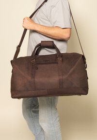 Brown leather duffel bag with a zippered pocket, sturdy handles, adjustable shoulder strap, and brass hardware accents. Textured finish.