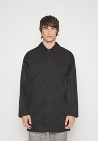 Abrigo negro impermeable de corte recto, con cierre de botones y bolsillos laterales. Presenta un cuello clásico y una textura lisa y mate.