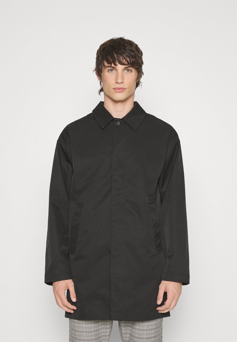 Abrigo negro impermeable de corte recto, con cierre de botones y bolsillos laterales. Presenta un cuello clásico y una textura lisa y mate.