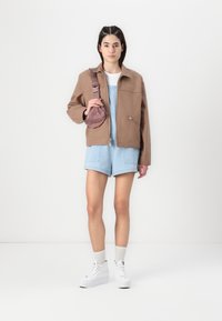 Hnědá oversized bunda, světlé denimové šortky, bílé vysoké tenisky a malá růžová taška. Hladký materiál a uvolněný střih.