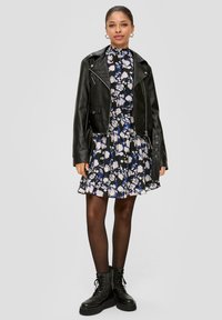 Zwarte leren cropped jas over een florale jurk met tinten blauw en wit, gecombineerd met zwarte veterschoenen.