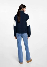Sweatshirt marine avec des accents blancs sur les épaules, col haut et poignets côtelés, associé à un jean flare bleu clair et des chaussures plateforme beiges.