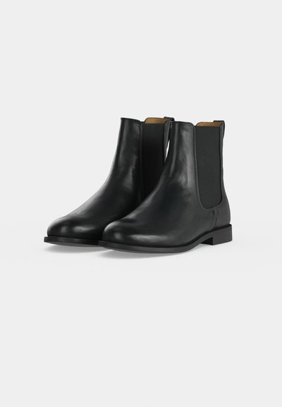 Paire de bottines Chelsea en cuir noir avec des panneaux latéraux élastiques et des petits talons sur un fond clair.