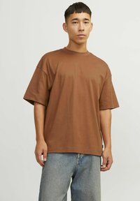 Hnědé oversized tričko vyrobené z měkké bavlny. Má krátké rukávy a kulatý výstřih. Kombinováno se světle modrými džíny.
