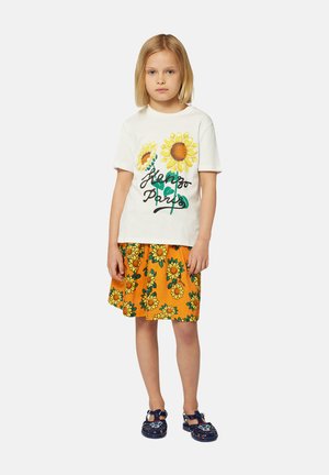 T-shirt en coton blanc avec un imprimé de tournesols jaunes et de feuilles vertes, associé à une jupe orange à motif de tournesols. Sandales bleues.