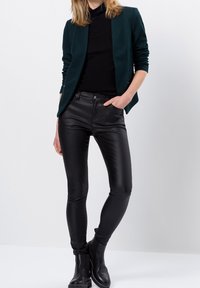 Jean skinny noir en cuir synthétique associé à un col roulé sombre et un blazer ouvert vert foncé. Des bottines noires complètent la tenue.