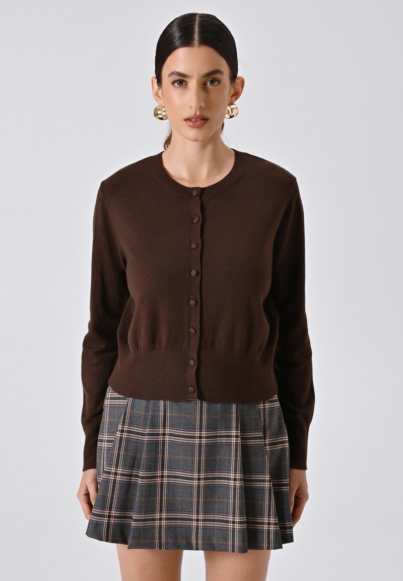 Cardigan marrone a maglia con scollo rotondo e chiusura frontale a bottoni, abbinato a una gonna a pieghe grigia a quadri con accenti rosa e blu.