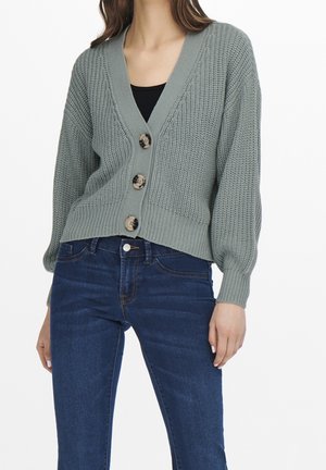 Femme portant un cardigan tricoté gris avec trois grands boutons et un jean bleu foncé, debout devant un fond blanc uni.