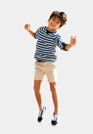 Blue and white striped long-sleeve shirt, beige shorts, navy sneakers. Boy jumps with arms raised, smiling against a plain background.