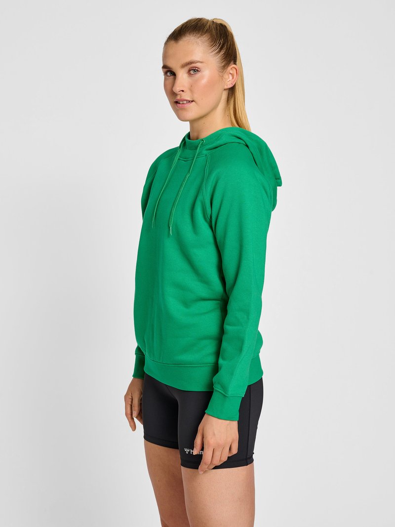 Jeune femme avec une queue de cheval blonde portant un sweat à capuche vert et un short de sport noir, debout devant un fond clair uni.