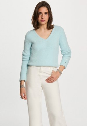 Femme portant un pull léger bleu en V avec des boutons sur les manches et un pantalon blanc taille haute, se tenant devant un fond uni.