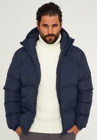 Marineblaue Steppjacke mit Kapuze und Reißverschluss, getragen über einem cremefarbenen Zopfmuster-Strickpullover. Glatte Textur, warmes Material.