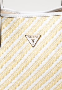 Sac en osier présentant un motif géométrique en crème et jaune pâle, avec une plaque en métal triangulaire sur le devant. Accents blancs lisses.