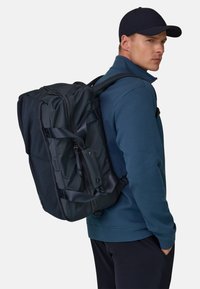 Zaino blu navy con più scomparti con cerniera, spallacci imbottiti e dettagli in rete, indossato sopra una felpa blu, dal design elegante.