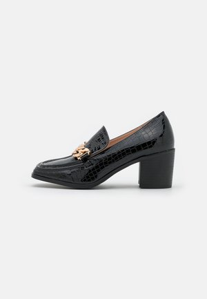 Mocassin noir à motif croco avec talon bloc et détail chaîne en or sur le dessus, présenté sur un fond blanc.