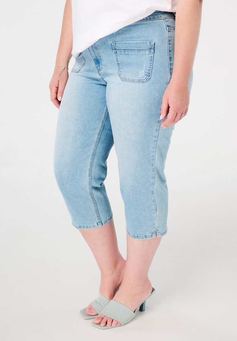 Pantalons capri en denim bleu clair avec une taille mi-haute, dotés de poches arrière et d'un ourlet cousu, accompagnés de chaussures ouvertes bleu clair.
