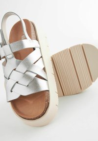 Hõbedased metallik sandaalid, millel on põimunud rihmad, kork jalaosa ja valge platvormi talla, millel on tekstuuritatud alune kanna paremaks haarde saavutamiseks.