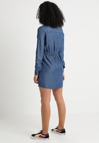 Denimklänning med urholkad midja, långa ärmar och en lätt A-linjeform. Bärs med svarta och röda sneakers.