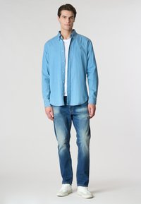 Camicia azzurra chiara a maniche lunghe con bottoni in denim, indossata sopra una maglietta bianca; abbinata a jeans blu sbiaditi e sneakers bianche.