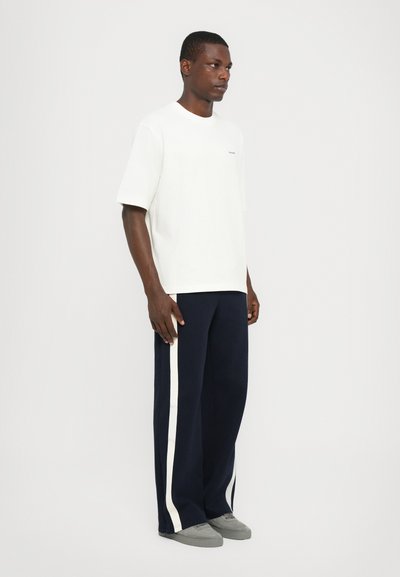Camiseta blanca de manga corta con cuello redondo, combinada con pantalones anchos de color azul marino oscuro que tienen rayas laterales blancas y zapatillas gris.
