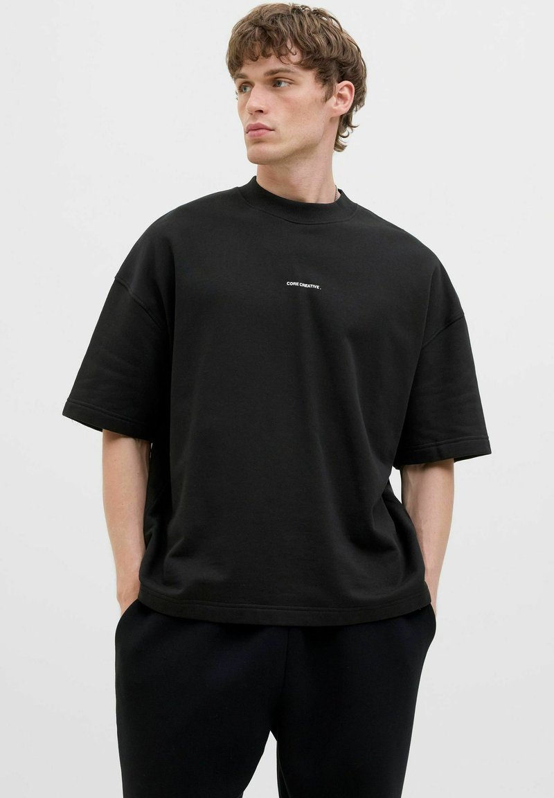 T-shirt noir oversize à manches courtes en coton avec un col rond, présentant un logo blanc "CORE CREATIVE" imprimé sur la poitrine.
