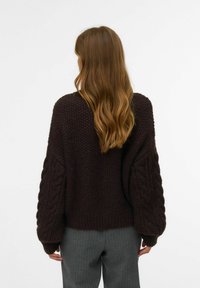 Pull en maille surdimensionné couleur marron foncé avec des manches en tricot torsadé, des poignets côtelés et un corps texturé, associé à un pantalon rayé gris.