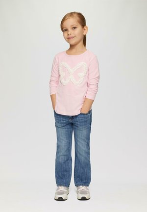 Maglietta a maniche lunghe rosa chiaro con un design di farfalla bianca texturizzata, abbinata a jeans in denim blu e sneakers grigie.