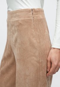 Pantalones de pana beige con textura acanalada vertical, que cuentan con una cremallera lateral y un acabado suave y aterciopelado.