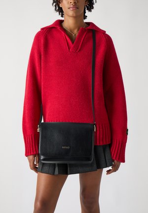 Maglione rosso a maglia con colletto, dotato di polsini a costine. Borsa a tracolla in pelle nera con patta e logo dorato, abbinata a una gonna scozzese.