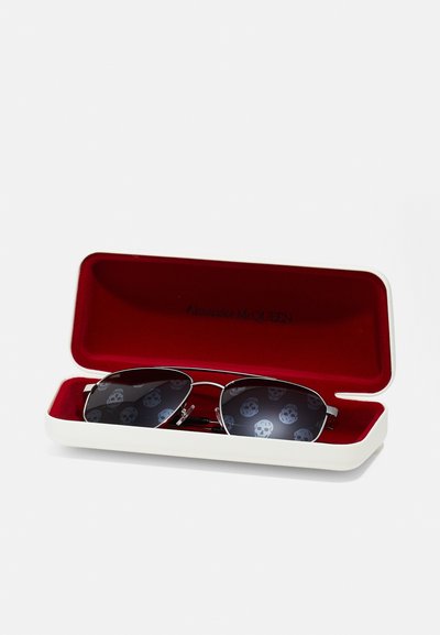Sunglasses in a white case with a red interior. Frames are silver metal, with dark lenses featuring a repeated skull pattern.