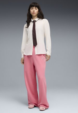 Frau trägt weißes Hemd mit Kragen und dunkler Krawatte, lockere rosa Hose und passende rosa Puma-Hausschuhe, steht vor einem grauen Verlaufshintergrund.