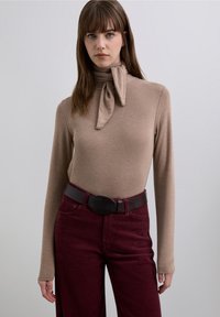 Top bej cu mâneci lungi și detaliu modelat cu panglică la gât, asortat cu pantaloni din catifea cu caneluri de culoare burgundy închis și o curea neagră cu cataramă ovală.
