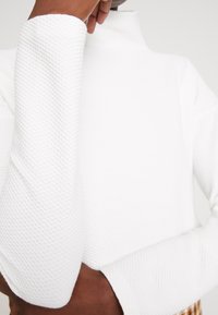 Top bianco a collo alto, strutturato, con motivo a nido d'ape e maniche leggermente svasate.