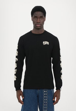 Black long-sleeve t-shirt with white logo on the chest and graphic designs on sleeves; paired with blue denim pants featuring studs.