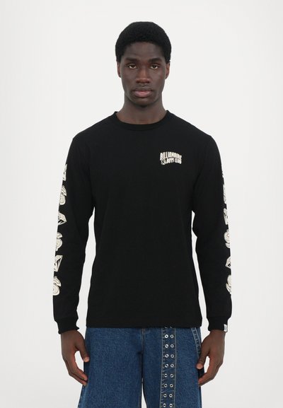 T-shirt noir à manches longues avec logo blanc sur la poitrine et designs graphiques sur les manches ; assorti à un pantalon en denim bleu avec des clous.