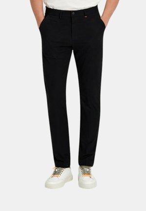 Uomo con pantaloni neri slim e sneakers bianche con lacci grigi e arancioni, in piedi con le mani in tasca.