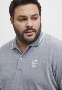 Szürke poloing, textúrált felülettel, fehér logóval és kontrasztos gallérral. Rövid ujjú és gombos záródású.