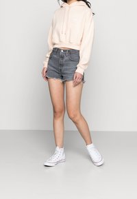 Hoodie corta rosa chiaro con tasca frontale, abbinato a pantaloni shorts in denim grigi a vita alta con orli sfrangiati e sneakers alte bianche.