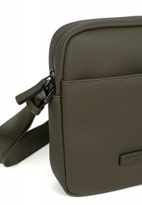 Borsa a tracolla rettangolare verde oliva in materiale sintetico testurizzato, con chiusura a zip e tracolla regolabile. Presenta una tasca frontale e dettagli con logo.