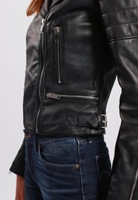 Giubbotto da motociclista in pelle nera con spalle trapuntate, numerous tasche con zip argentate e una cinghia regolabile con fibbia sul lato.