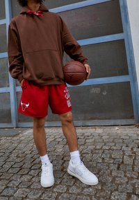 Brun zip-hoodie, röda basketshorts med Chicago Bulls grafik, vita sneakers och en basketboll i handen. Texturerad pavébakgrund.