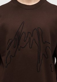 Καφέ πουλόβερ με στρογγυλή λαιμόκοψη και αφηρημένο μαύρο σχέδιο γραμμής στο στήθος, φορεμένο από άτομο με ορατό λαιμό και ώμους.