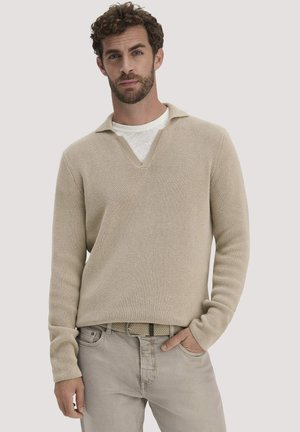 Mann mit lockigem Haar, der einen beigen V-Ausschnitt-Pullover über einem weißen Hemd und beigen Hosen trägt, steht mit einer Hand in der Tasche.