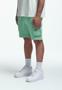 Hellgrüne Sweatshorts mit einem Kordelzug, ausgestattet mit einer Seitentasche. Kombiniert mit weißen Sneakers und Crew-Socken. Oben ein schlichtes weißes T-Shirt.