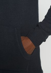 Maglione blu scuro con texture a coste, dotato di tasca frontale e polsini aderenti. Il design include un col a V per un tocco di eleganza.