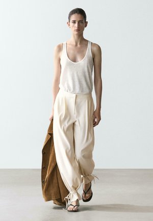 Femme marchant vers l'avant, portant un haut beige sans manches, un pantalon ample crème noué aux chevilles, des sandales noires, tenant une veste marron dans sa main droite.