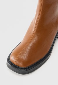 Stivale alla caviglia in pelle marrone con una superficie liscia, punta quadrata e suola in gomma nera. Presenta cuciture lungo il lato e un design elegante.