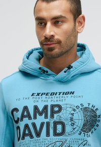 Man with trimmed beard wearing blue hoodie with black text and map graphics related to northern polar expedition.